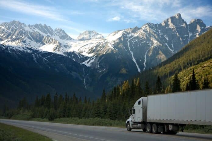 A semi-truck travels along a highway with snow-capped mountains